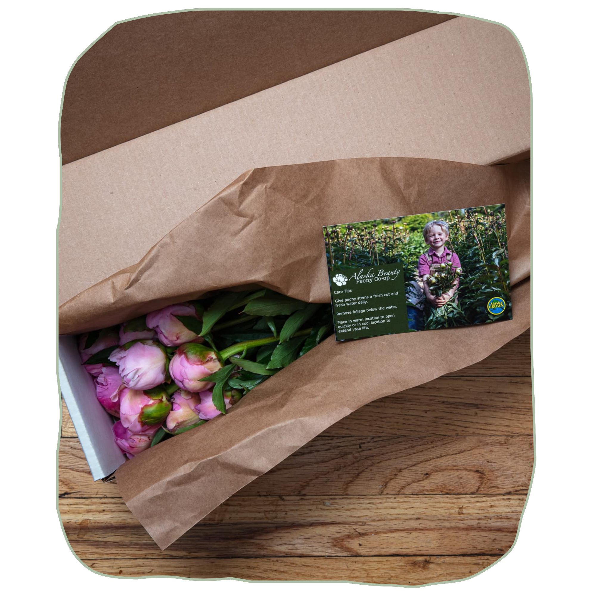 Bouquet of pink flowers wrapped in brown paper with a business card on a wooden surface | Alaska Beauty Peony Cooperative | Homer, AK